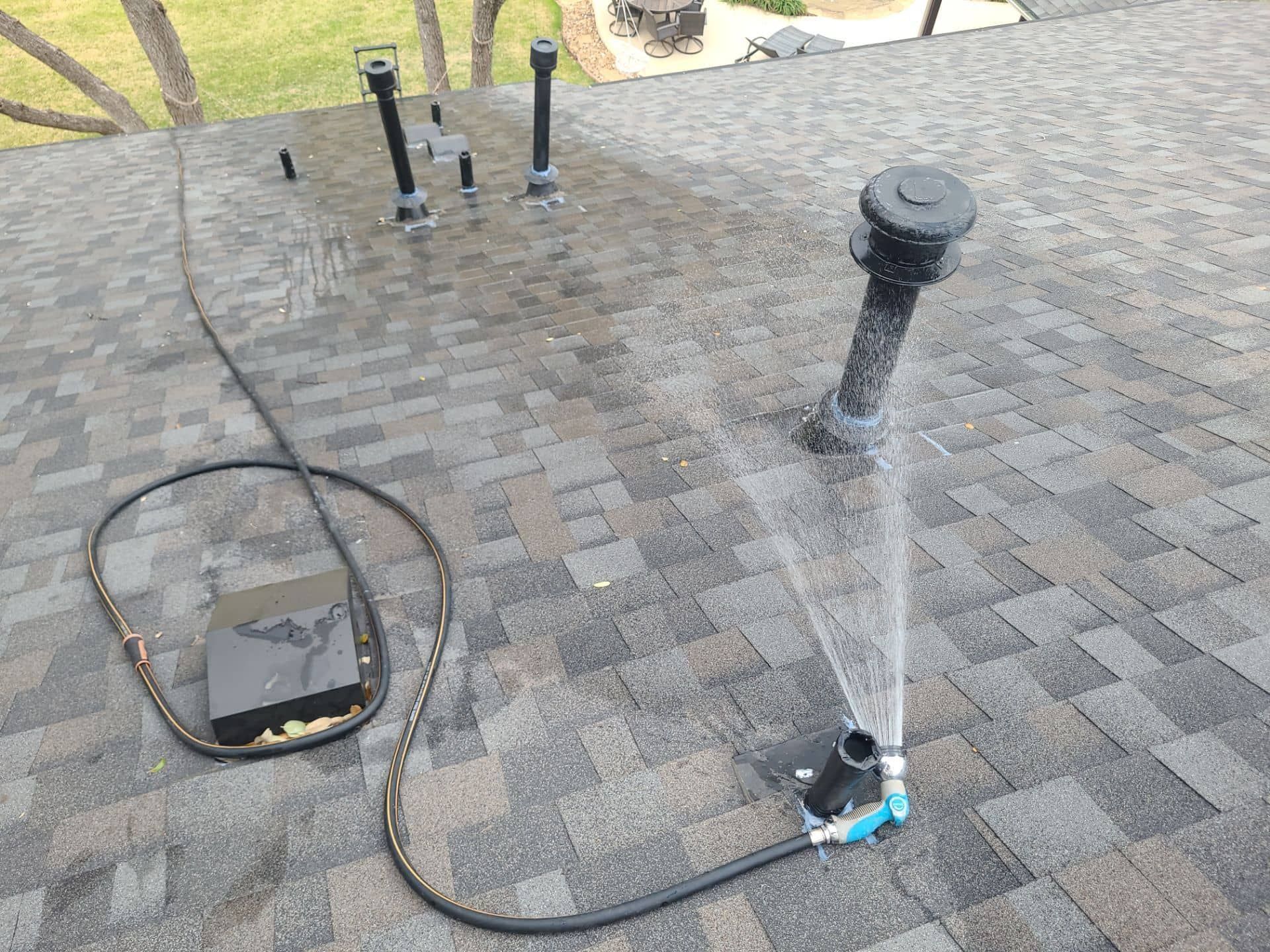 Water spraying on a shingled roof from a black hose connected to a device; several pipes nearby.
