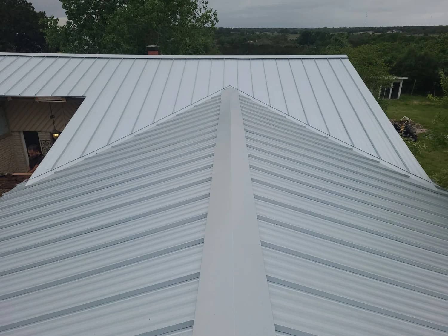 Metal roof on a building, angled view showing the ridgeline and panels of the roof with a light, overcast sky in the background.