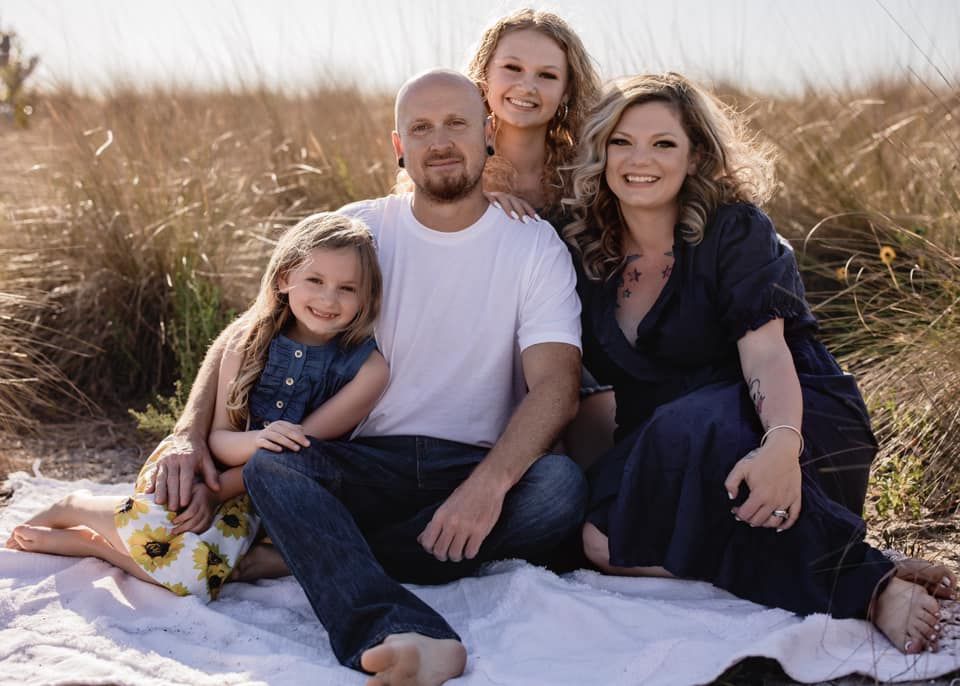 Family of four sitting on a beach blanket: man, woman, and two daughters smiling.