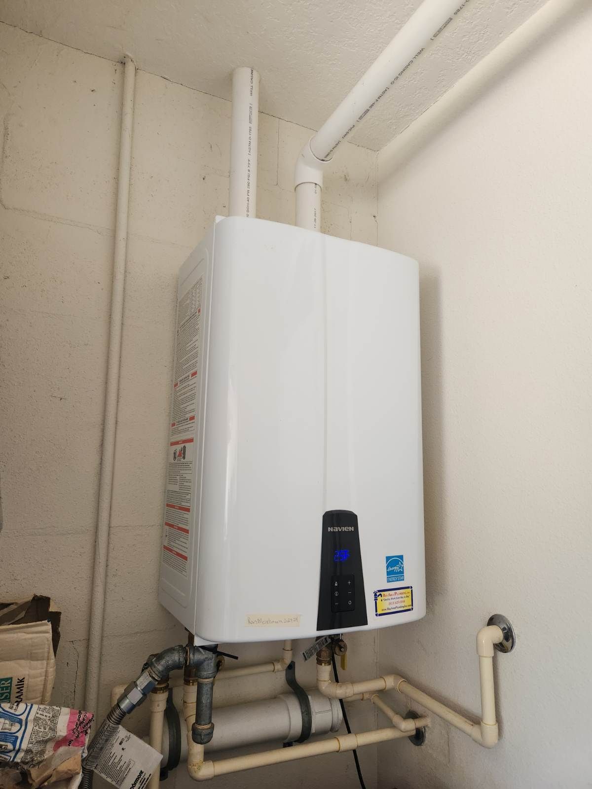 A white tankless water heater mounted on a wall in a utility room with attached pipes.