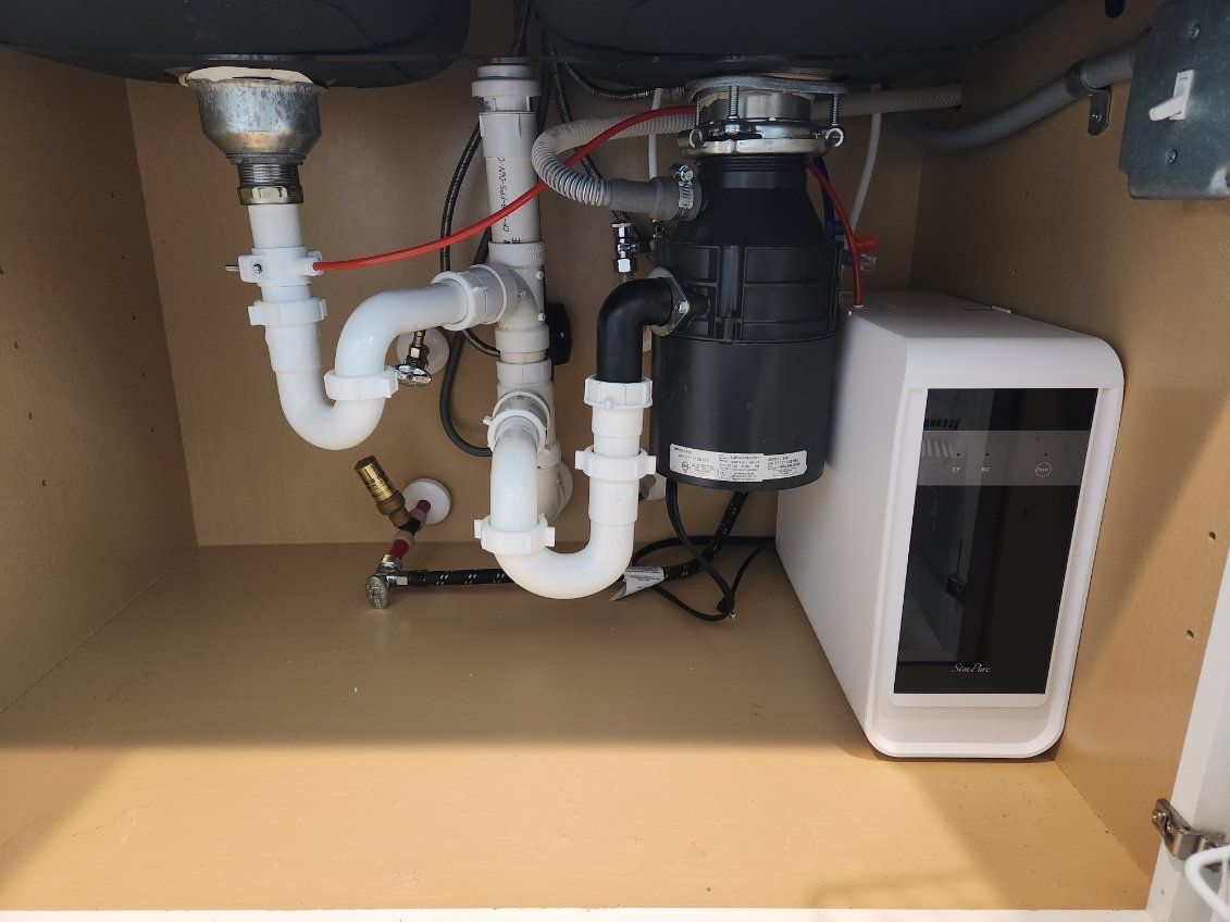 Undersink plumbing: White pipes, garbage disposal, water filter in a wooden cabinet.