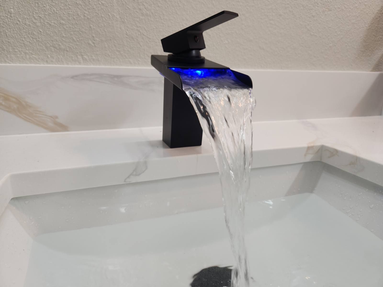 Black waterfall faucet with blue LED over a white sink, water flowing.