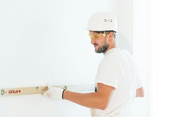 A man wearing a hard hat and goggles is measuring a wall with a level.