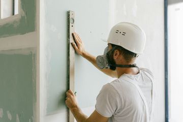A man wearing a helmet and a gas mask is measuring a wall with a level.