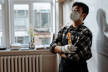 A man wearing a mask and gloves is standing in a room with his arms crossed.