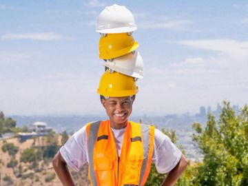 A construction worker is holding a stack of hard hats on his head.