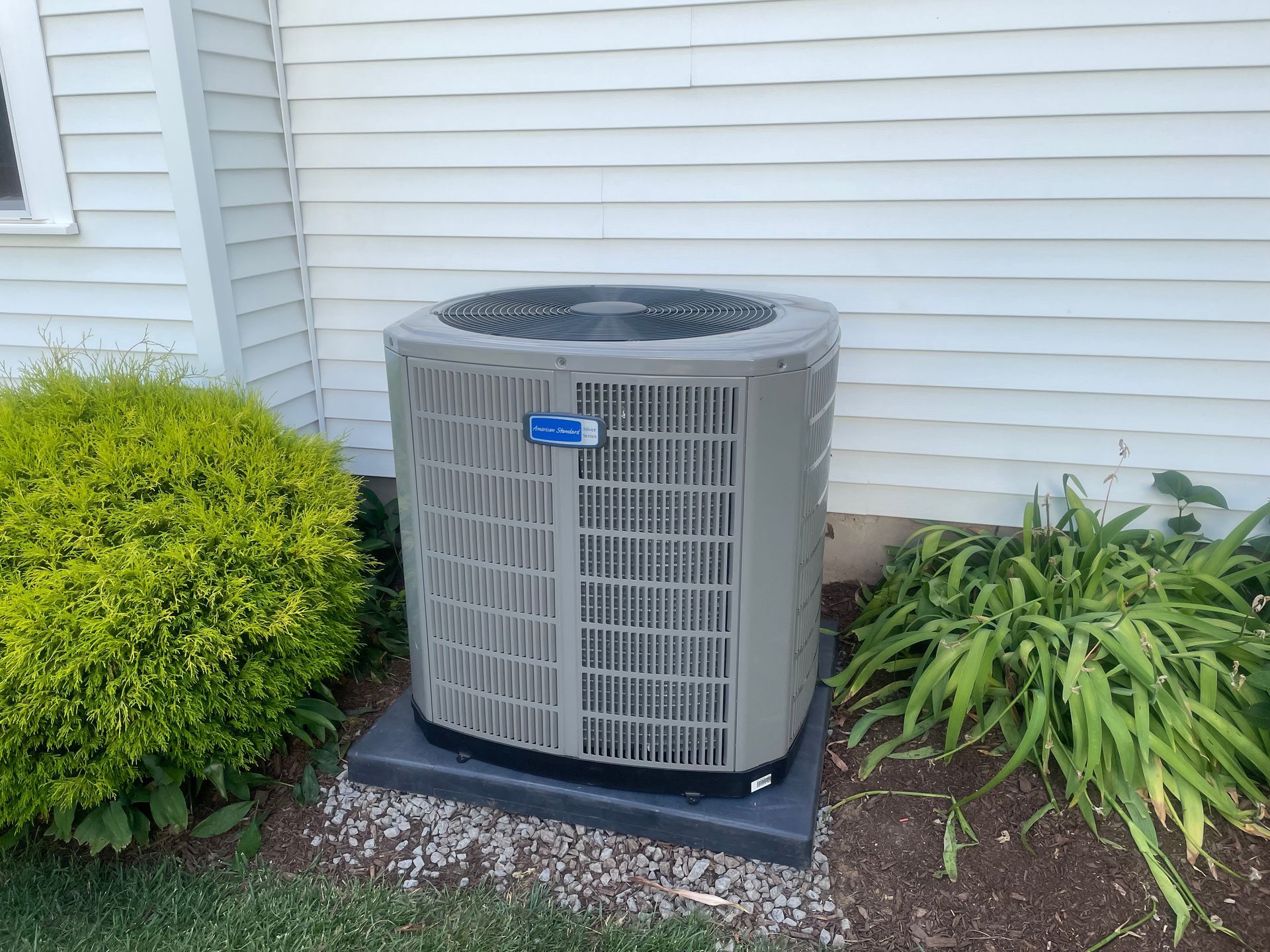 An air conditioner is sitting outside of a house next to a bush.
