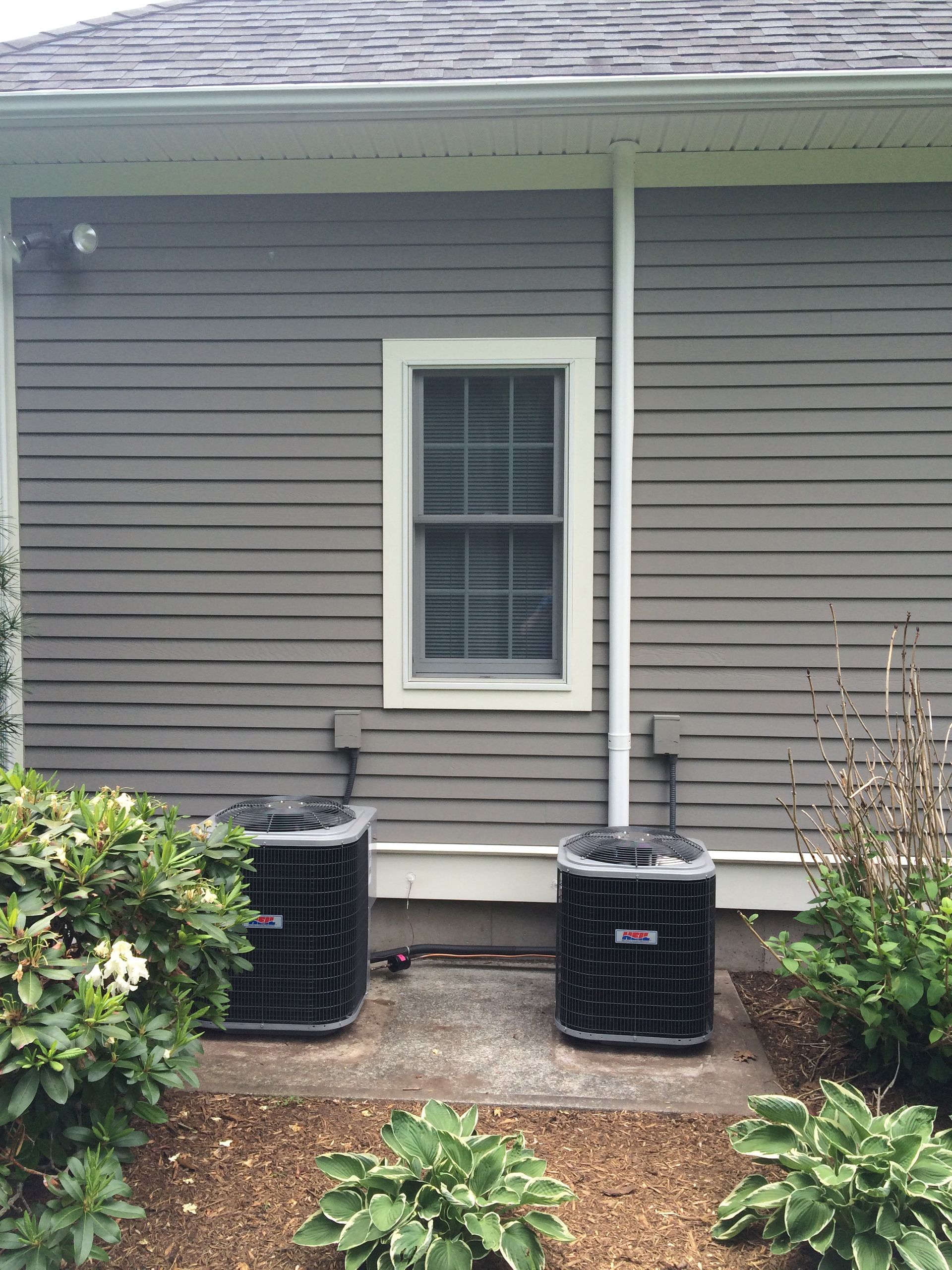 Two air conditioners are sitting outside of a house next to a window.