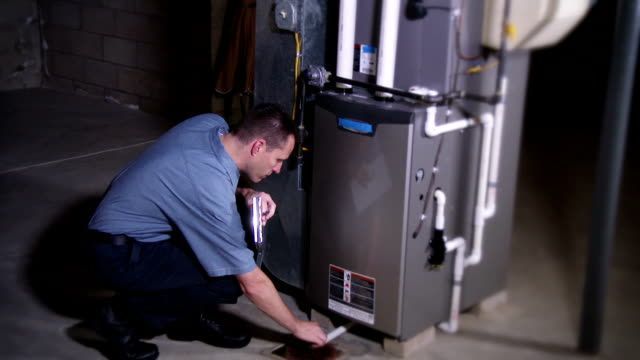 A man is kneeling down in front of a furnace in a basement.