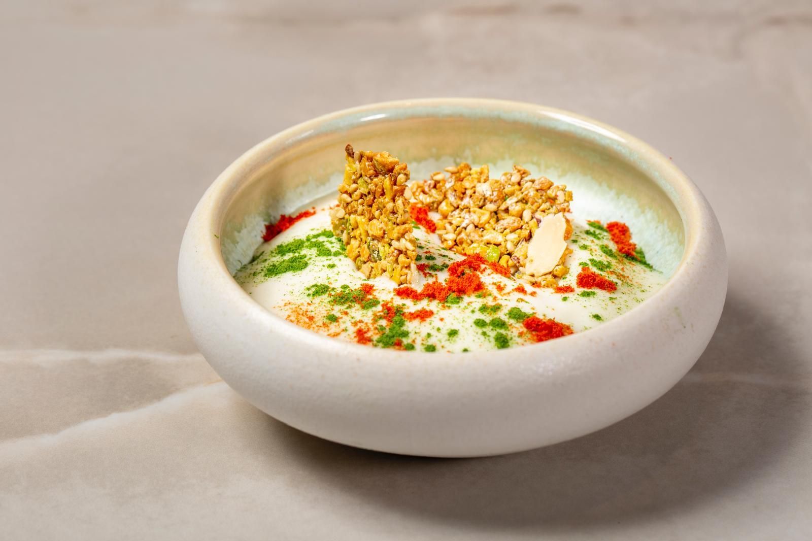 Una ciotola di ceramica bianca riempita con una crema, guarnita con granola, spezie rosse ed erbe aromatiche fresche.