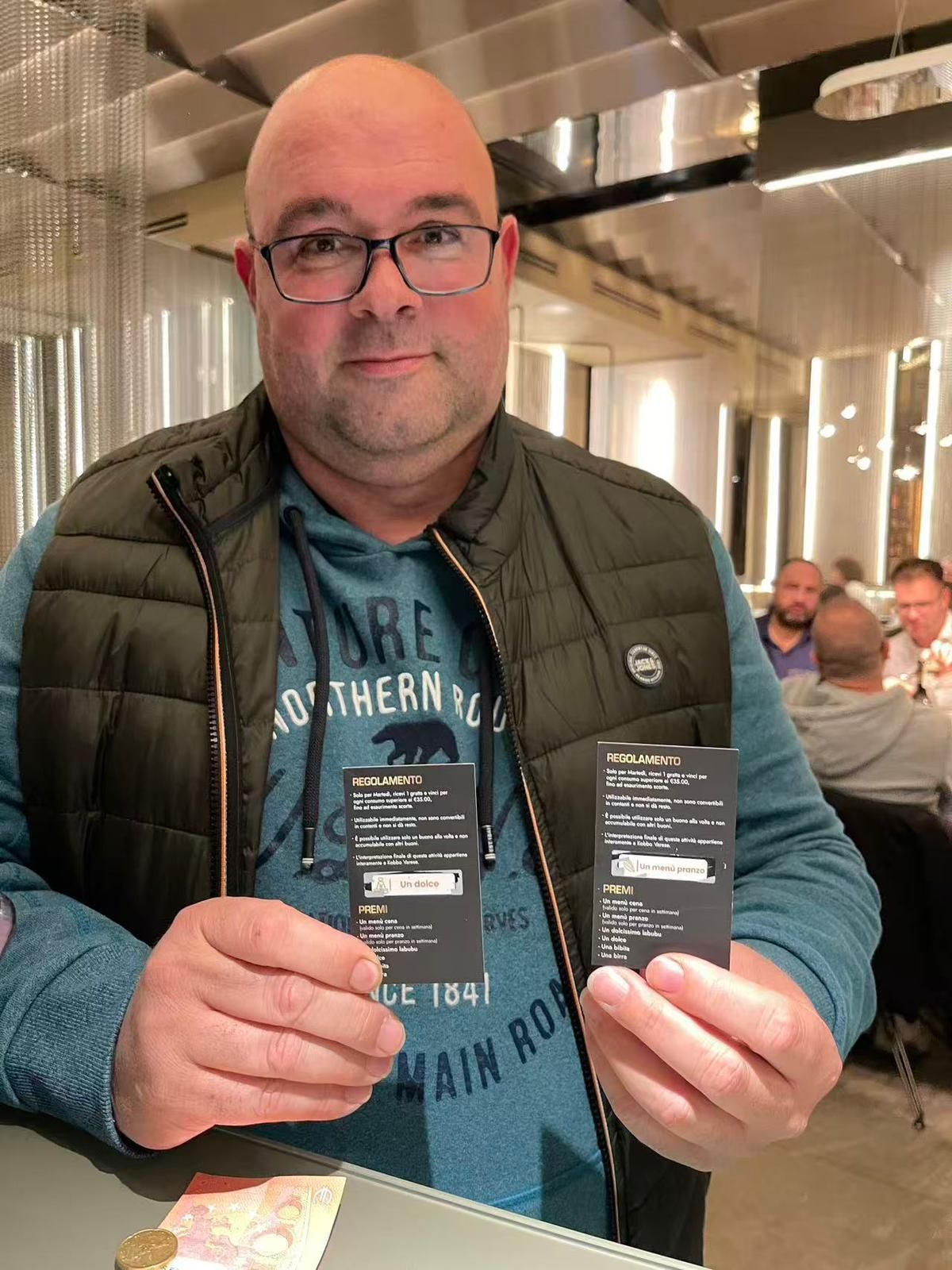 In un ristorante, un uomo con occhiali e gilet tiene in mano due piccole carte nere.