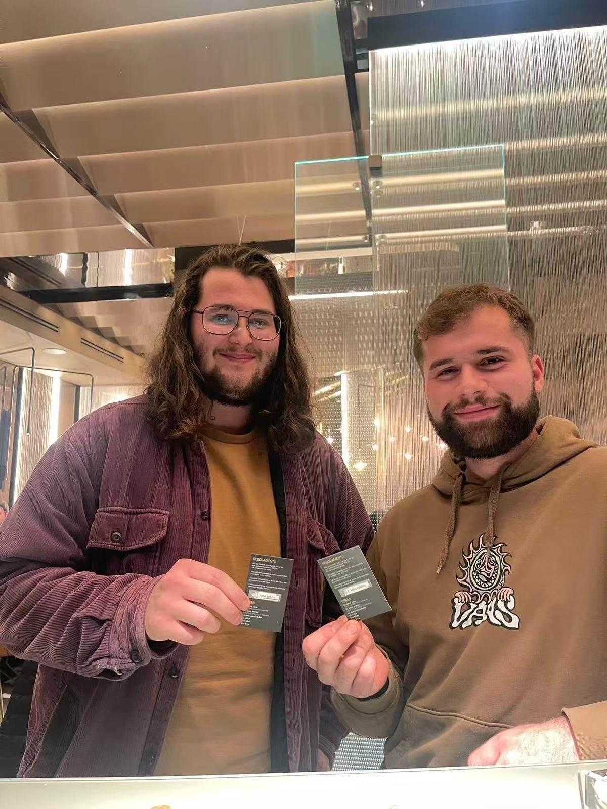 Due persone in abiti casual mostrano, sorridendo, dei piccoli cartoncini rettangolari scuri all'interno di un edificio.