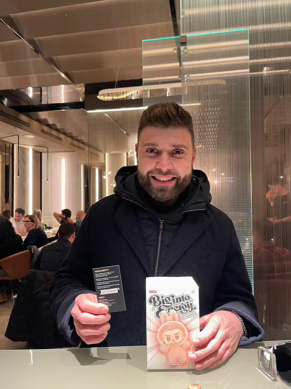 Una persona seduta in un ristorante tiene in mano un piccolo cartoncino nero e una scatola bianca con un'illustrazione.