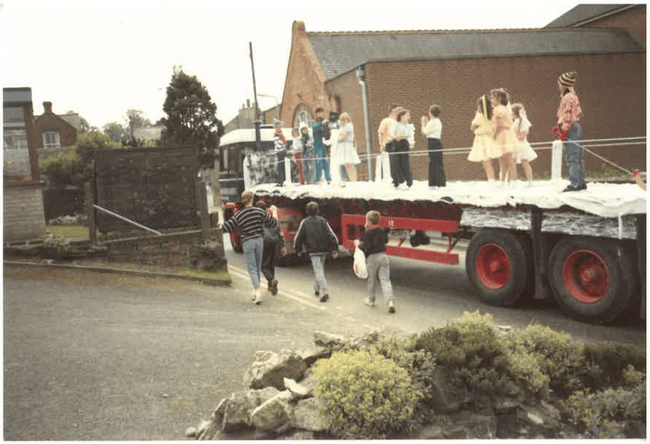 People run towards a flatbed truck carrying performers in ashby carnival parade. Brick buildings in the background. Old Ashby de la zouch 