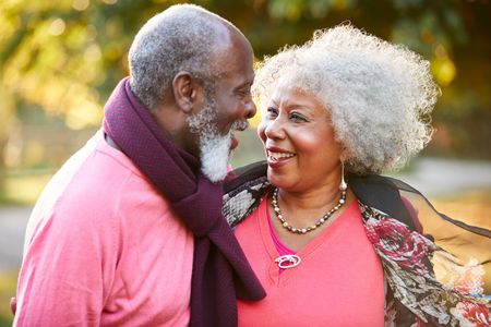 An older couple laughing together outdoors in the autumn, wearing warm clothing like sweaters and scarves.