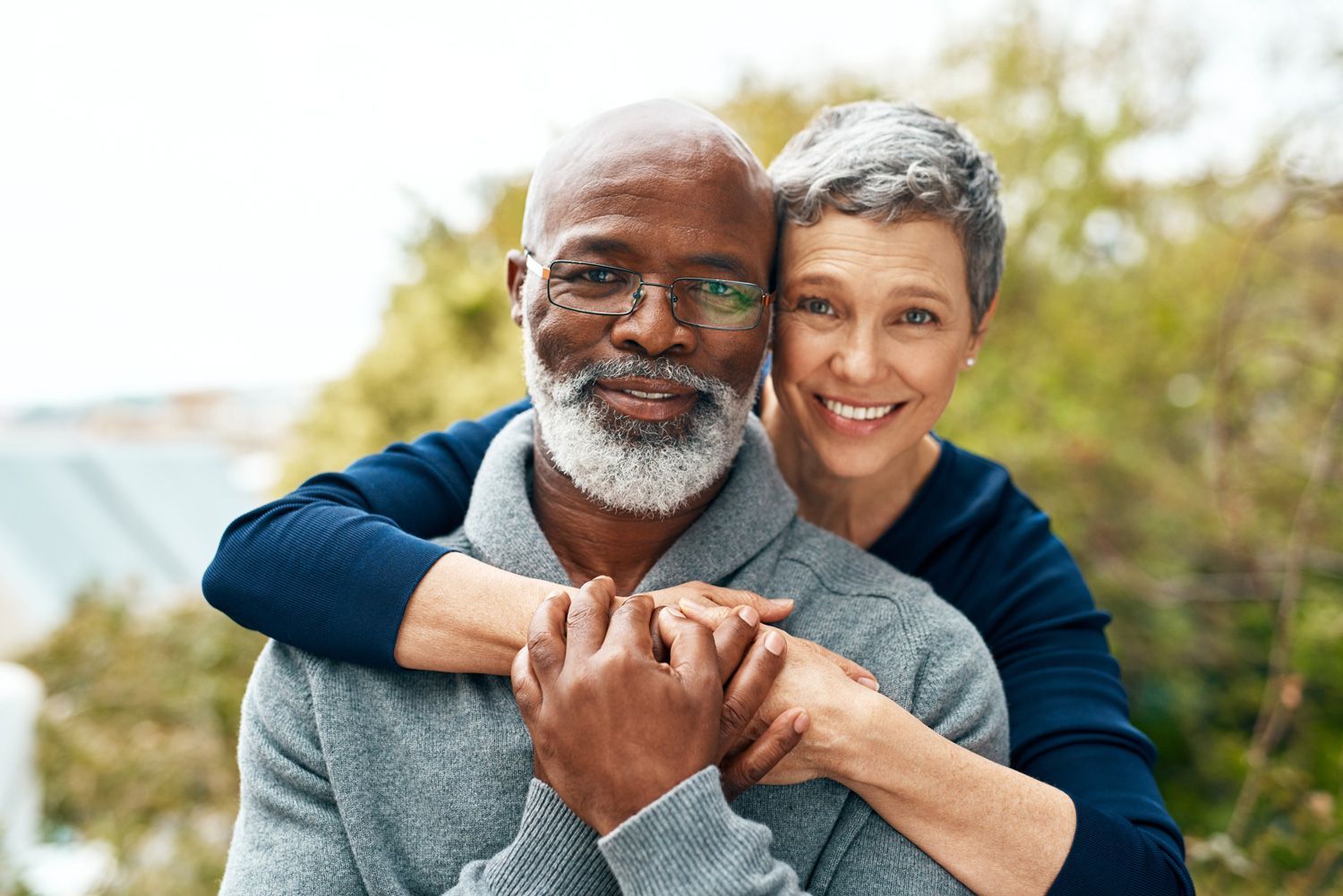 Two people smiling while one embraces the other from behind, set against a blurred outdoor background.