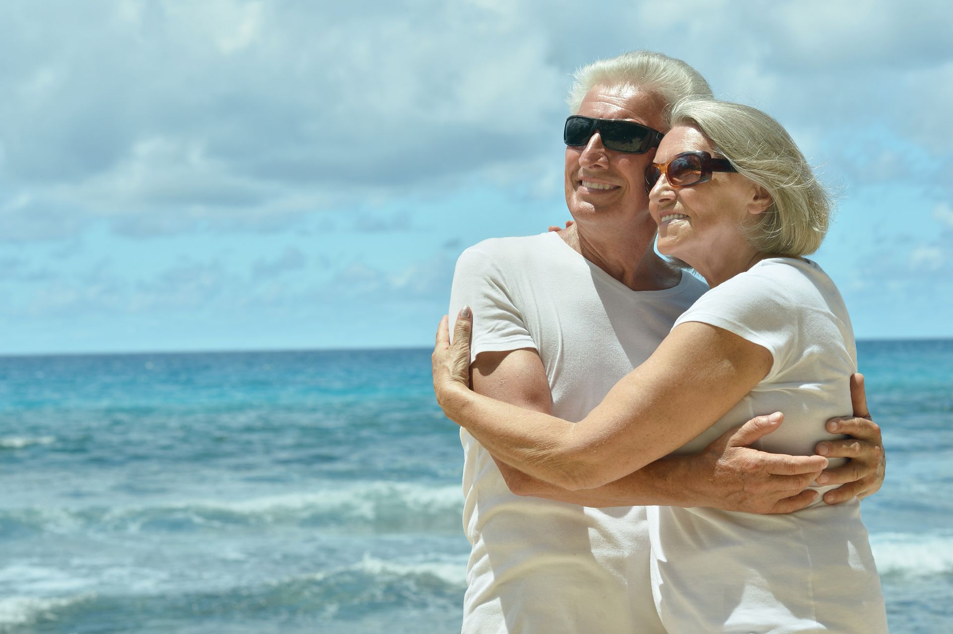 A pair wearing sunglasses and white shirts embrace while standing on a beach with the ocean in the background.