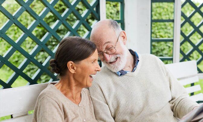 A couple laughs while sitting together on a white bench in front of a green lattice trellis.