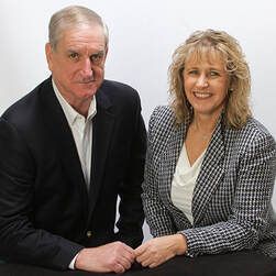 Two people in business attire sit side-by-side against a white background, holding hands and smiling at the camera.