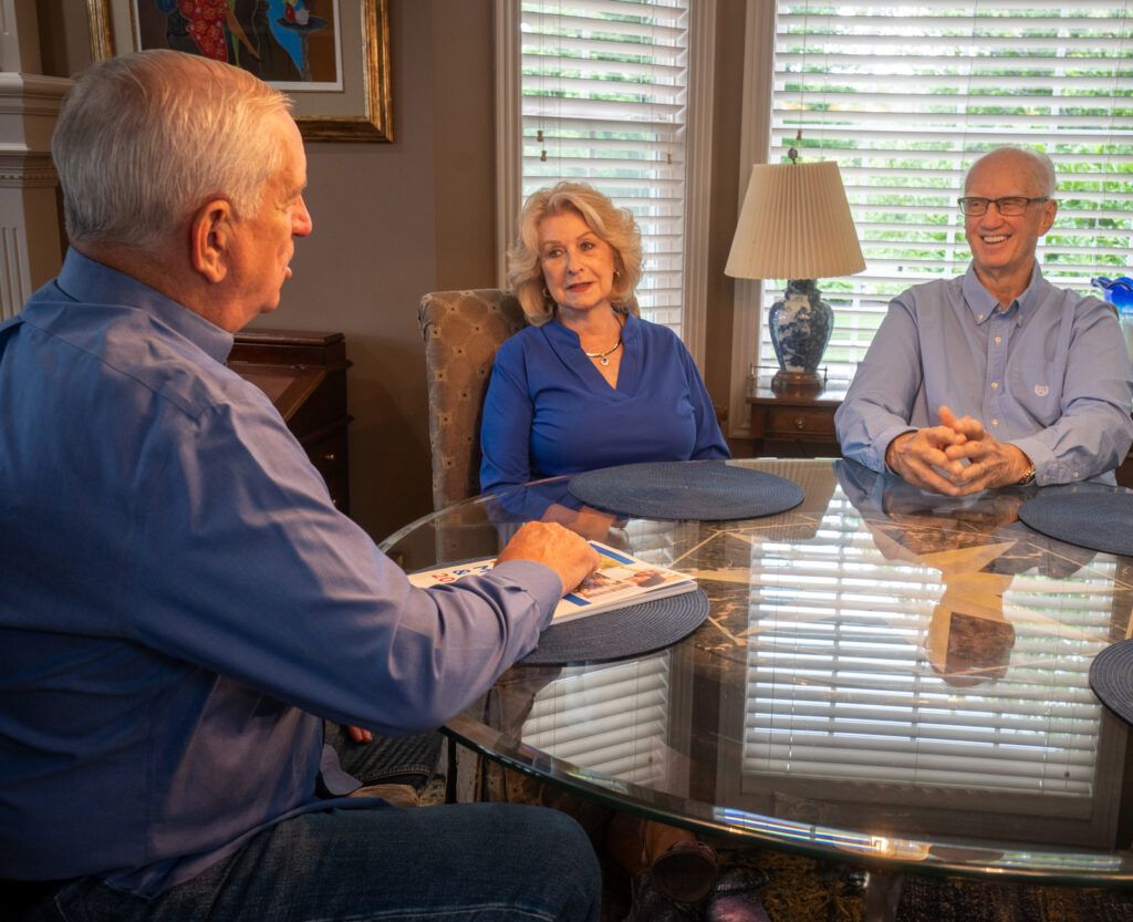 Three people in blue shirts are sitting around a circular glass dining table, engaged in a conversation.