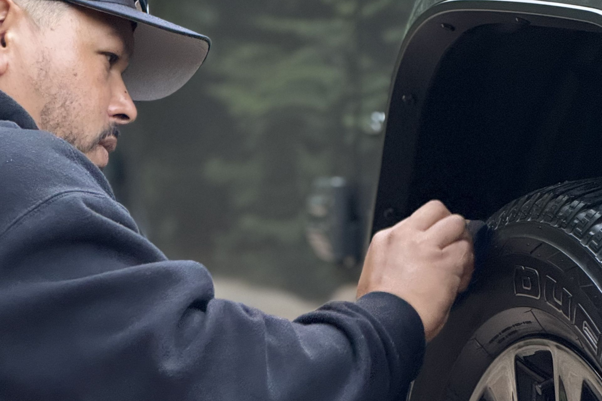 Man cleaning the tire of a vehicle, outdoors. He wears a hat and dark sweatshirt.