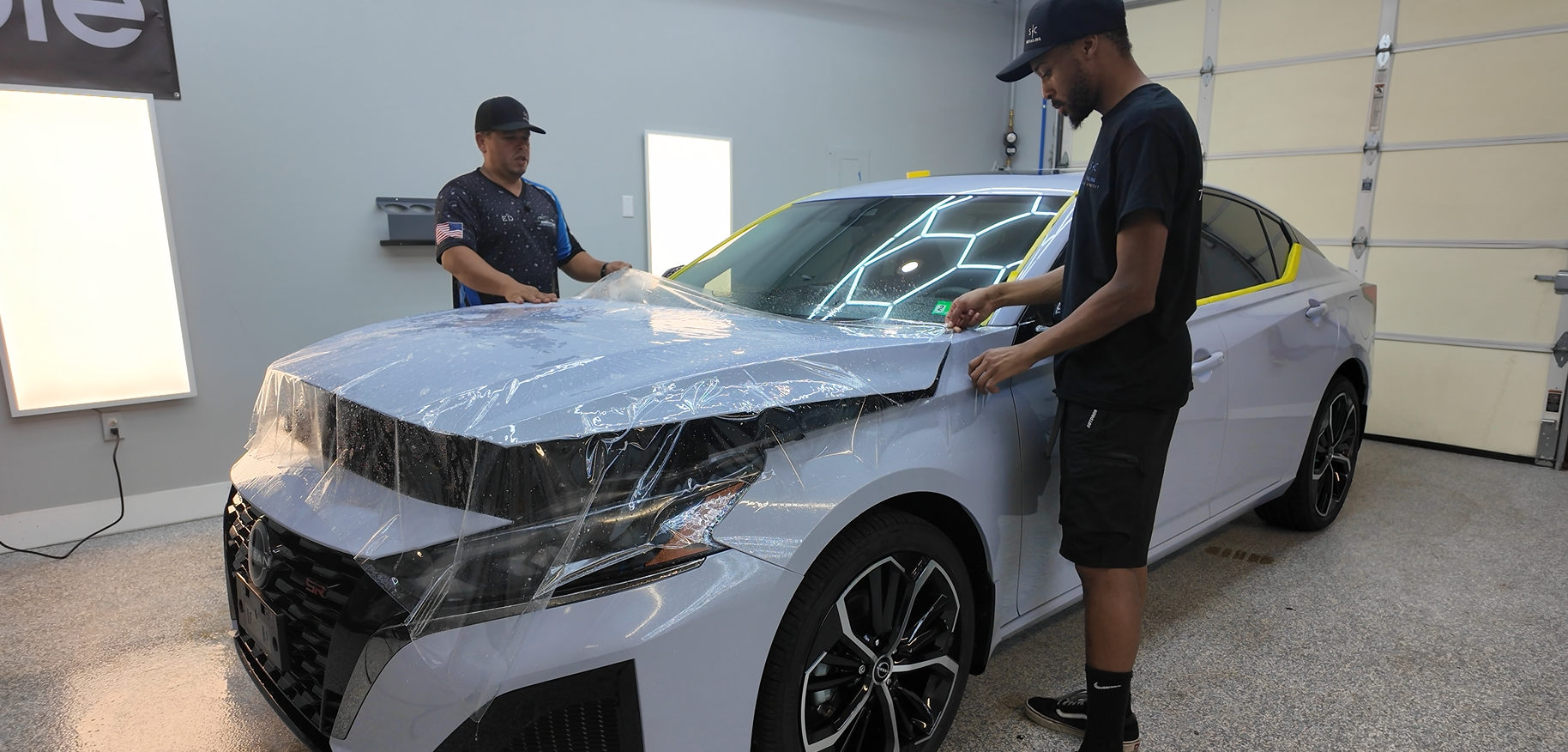 Two people applying protective film to the hood of a gray car in a garage.