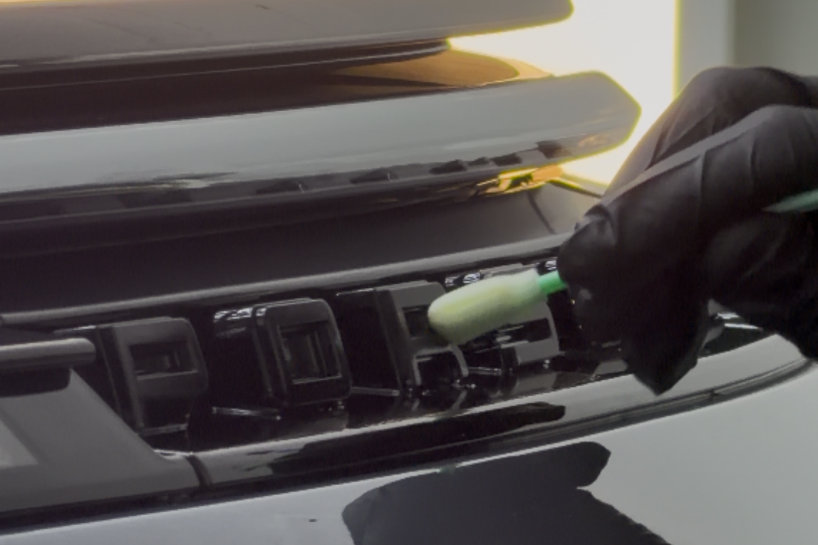 Gloved hand cleaning a car's black grill with a Q-tip.