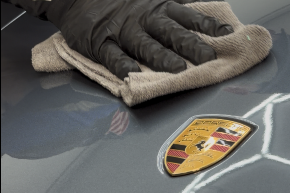 Gloved hand polishing a grey car surface with a cloth, next to a Porsche emblem.