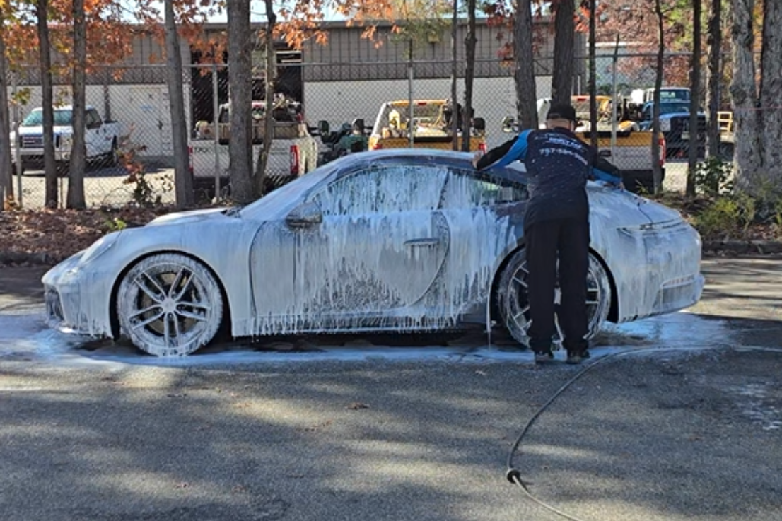 A white sports car covered in soap, being washed by a person in a blue shirt and black pants outside.