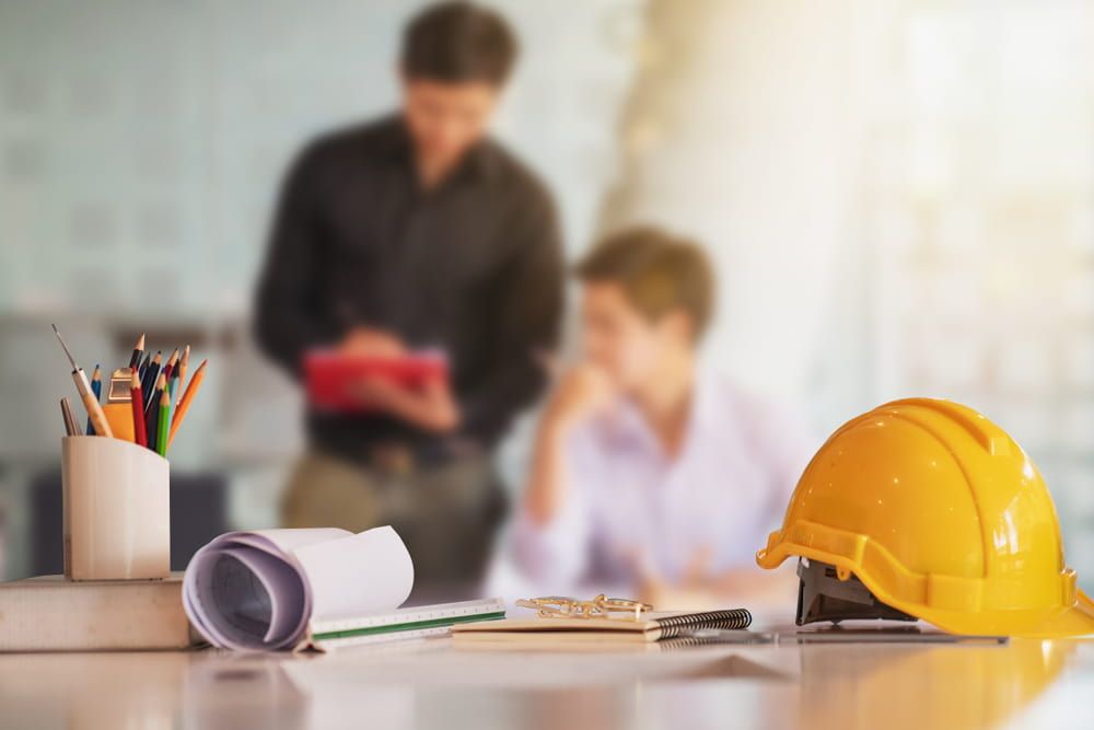 Yellow hard hat on a desk with blueprints and pencils; two blurred figures in the background discussing plans.