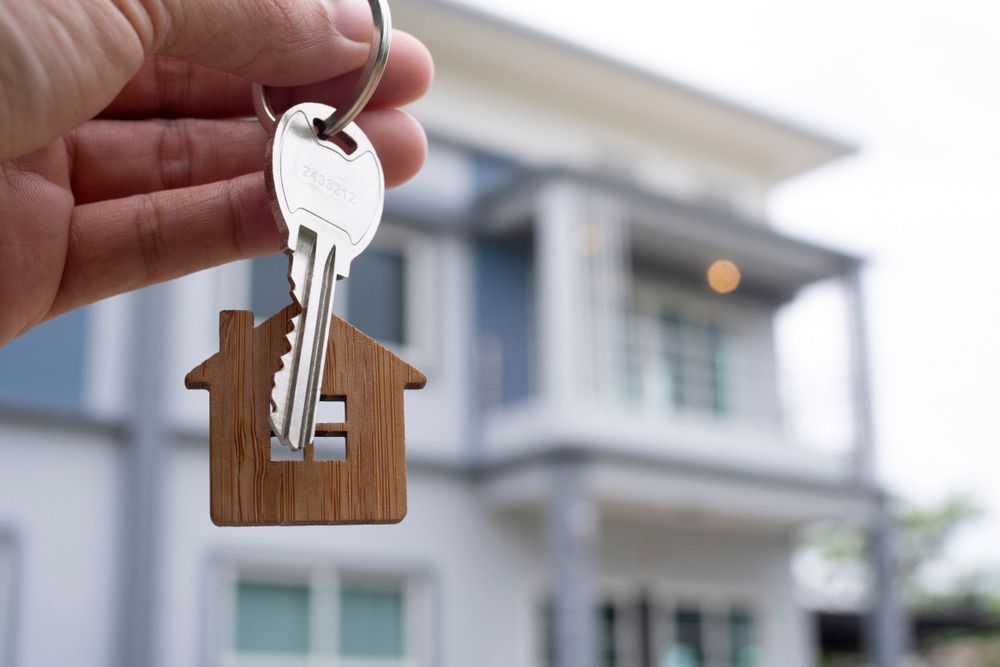 Hand holding house key with house-shaped keychain in front of a modern house.