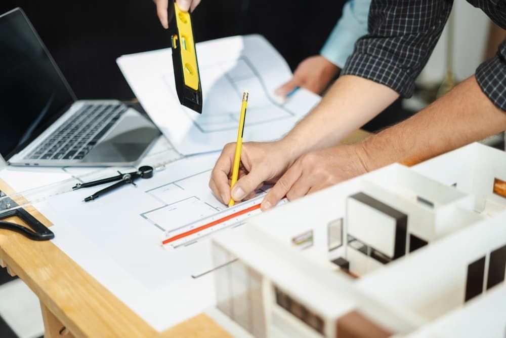 Architects working on blueprints, with a model house and laptop on a wooden desk.