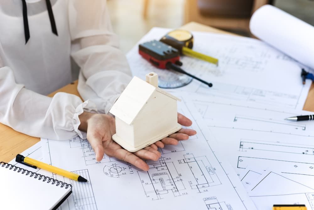Architect holding a model house, surrounded by blueprints, measuring tape, and a notepad.