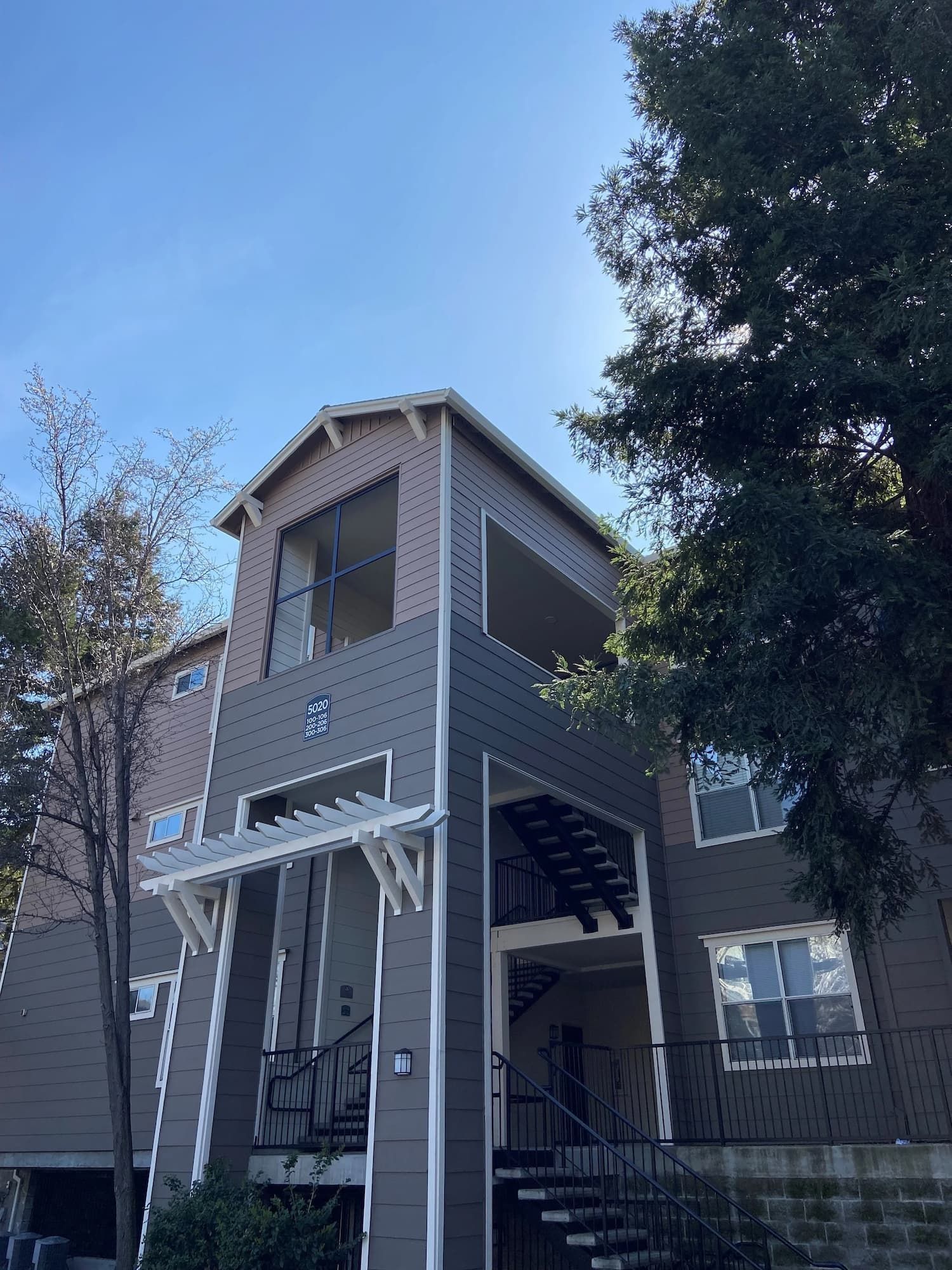 Multi-story residential building with gray and tan siding, trees, and a clear blue sky.