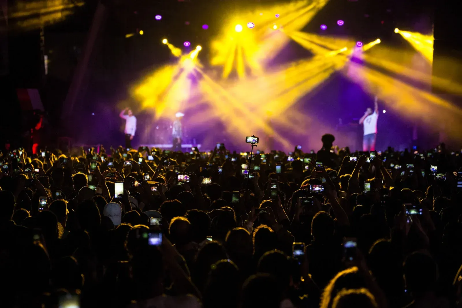 Concert crowd with raised phones, performers on stage under bright yellow and purple lights.