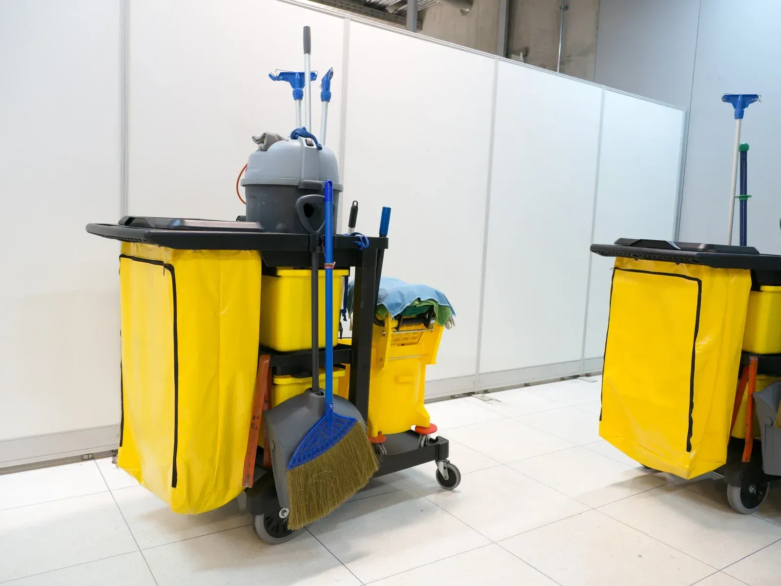Two cleaning carts with yellow bags, mops, and brooms on a white tiled floor.