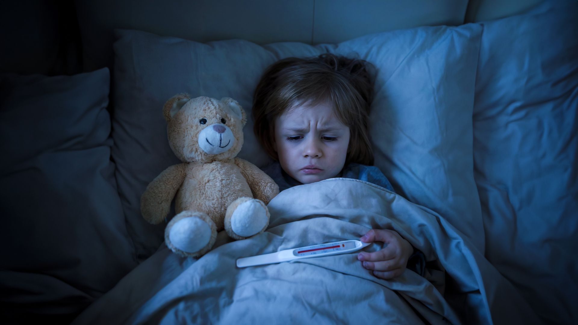 A little girl is laying in bed with a teddy bear and a thermometer.