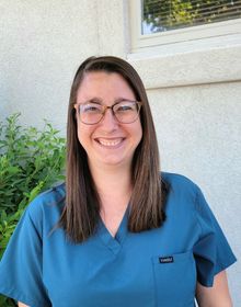 A woman wearing glasses and a blue scrub top is smiling for the camera.