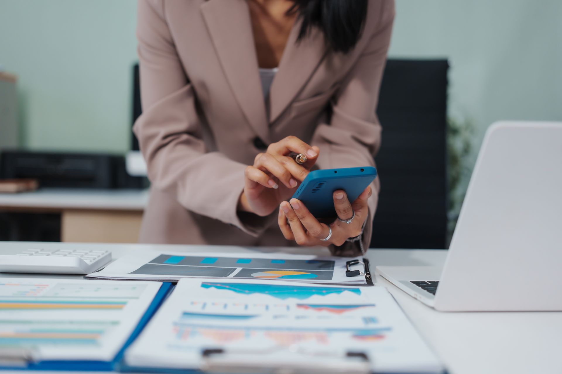 Woman in a blazer using a phone, working at a desk with charts and a laptop.