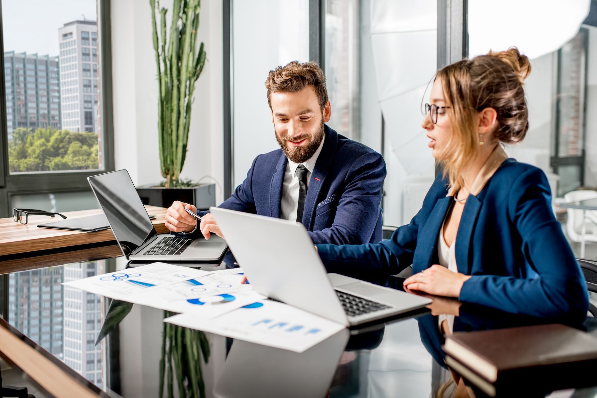 Two people in business attire reviewing charts on a laptop at a modern office desk.