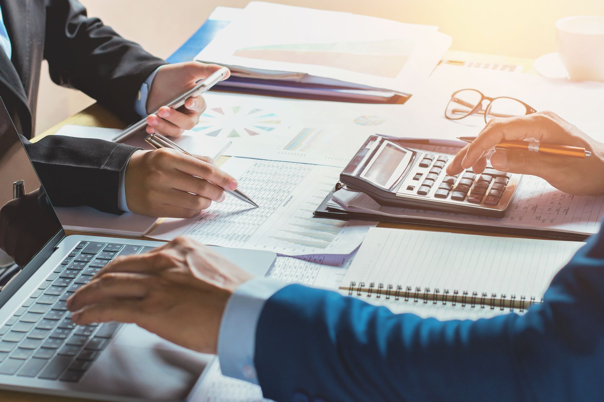 Two people reviewing financial documents, using a calculator and laptop, lit by sunlight.