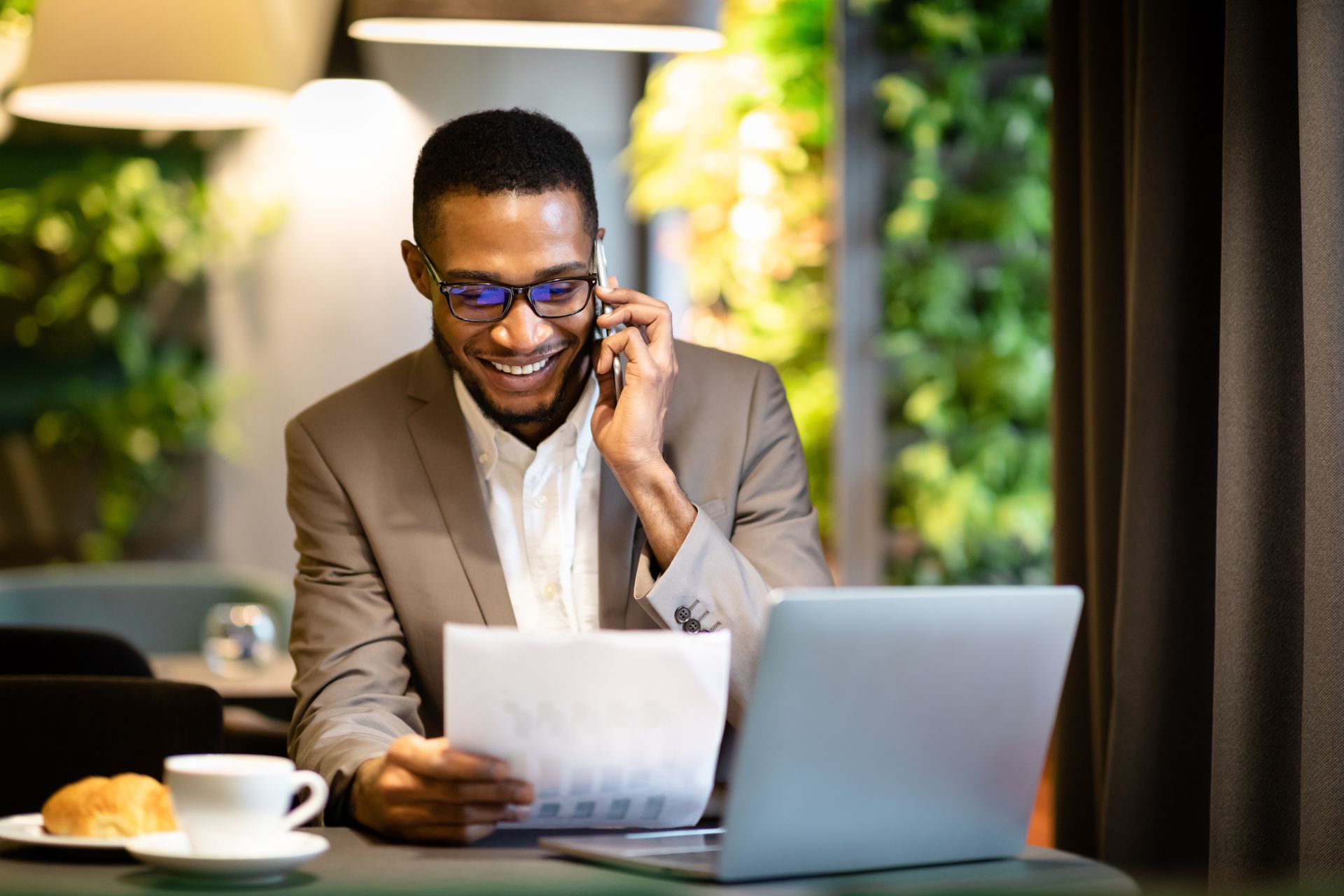 Man in suit on phone, smiling while reviewing papers, laptop open, sitting at table with coffee and pastry.