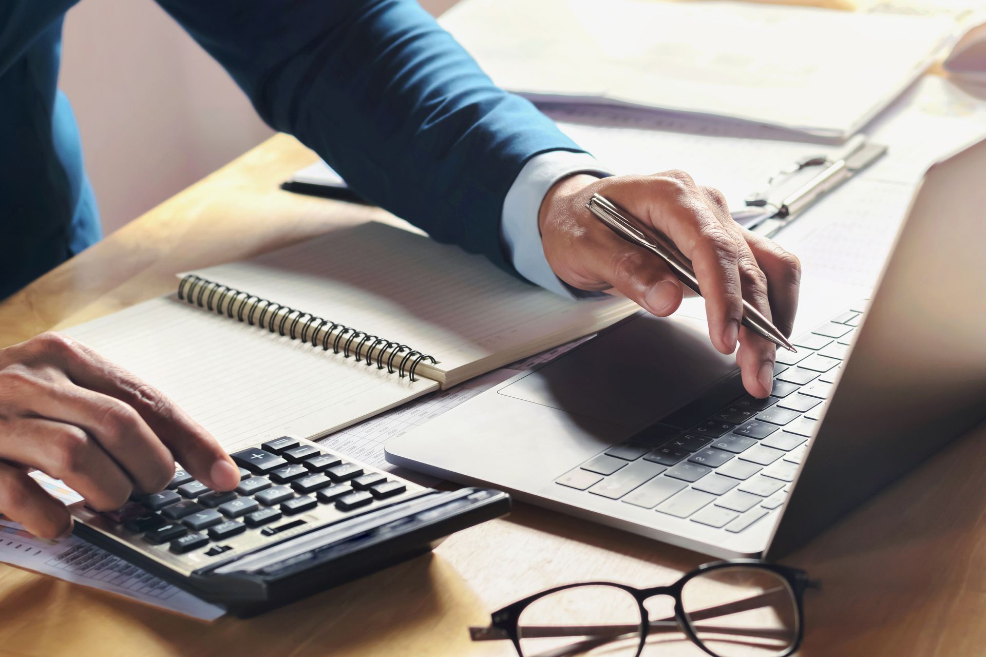 Person in a suit calculating with a calculator and laptop at a desk with notebook and glasses.