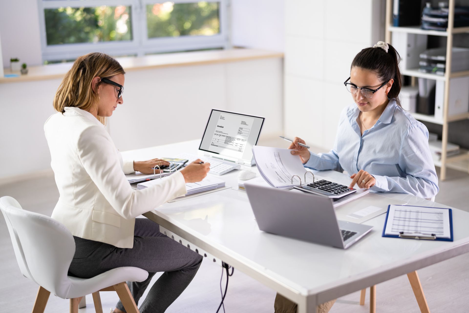 Two women working at a white desk with calculators, paperwork, and laptops in an office.