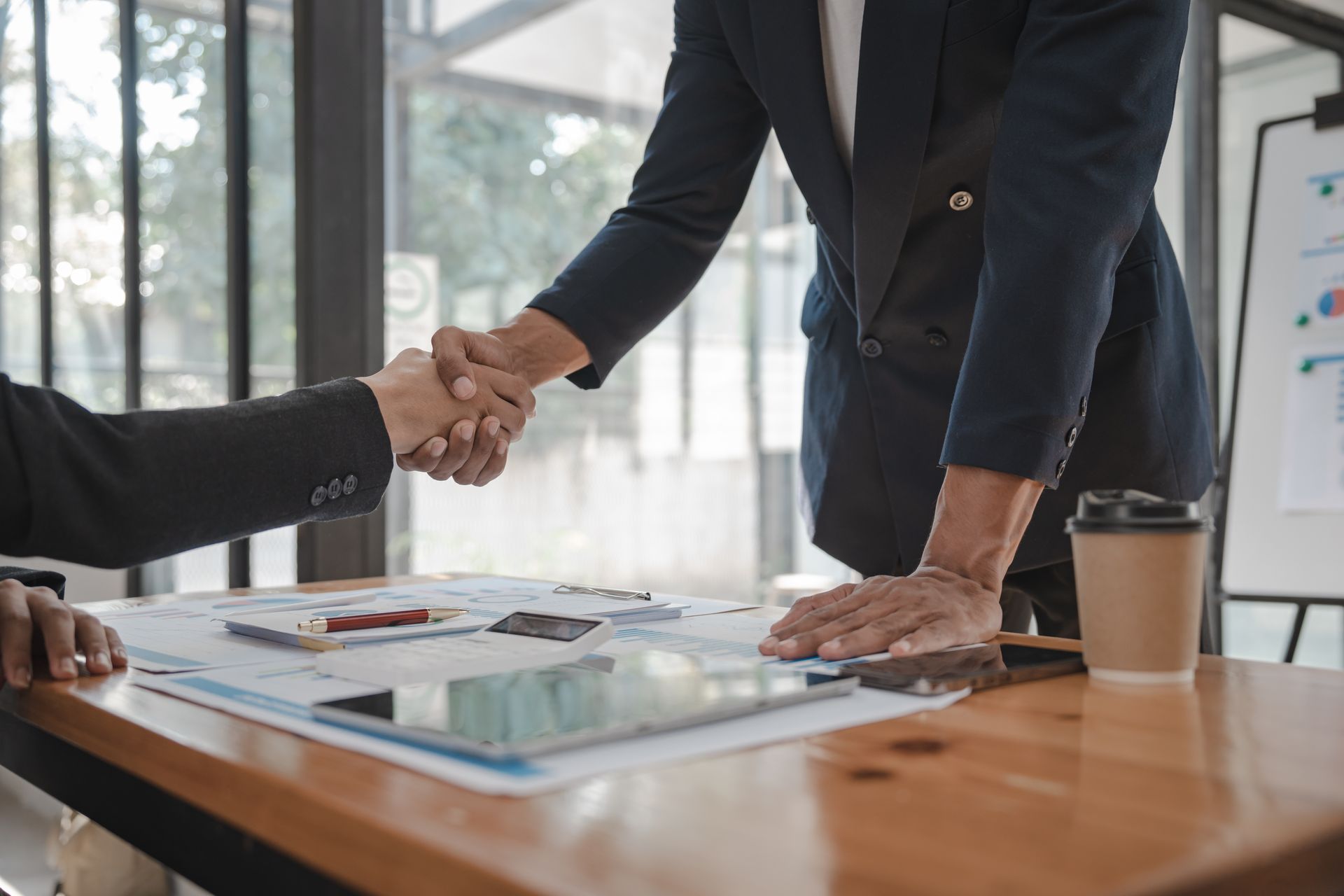 Two people in suits shaking hands over a table with papers, a tablet, and a coffee cup.