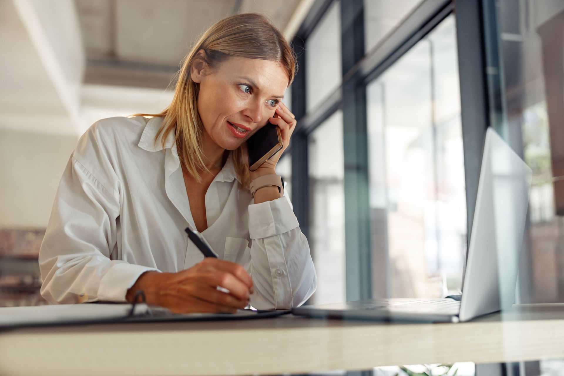 Woman on phone, writing notes, working at desk with laptop in front of a window.