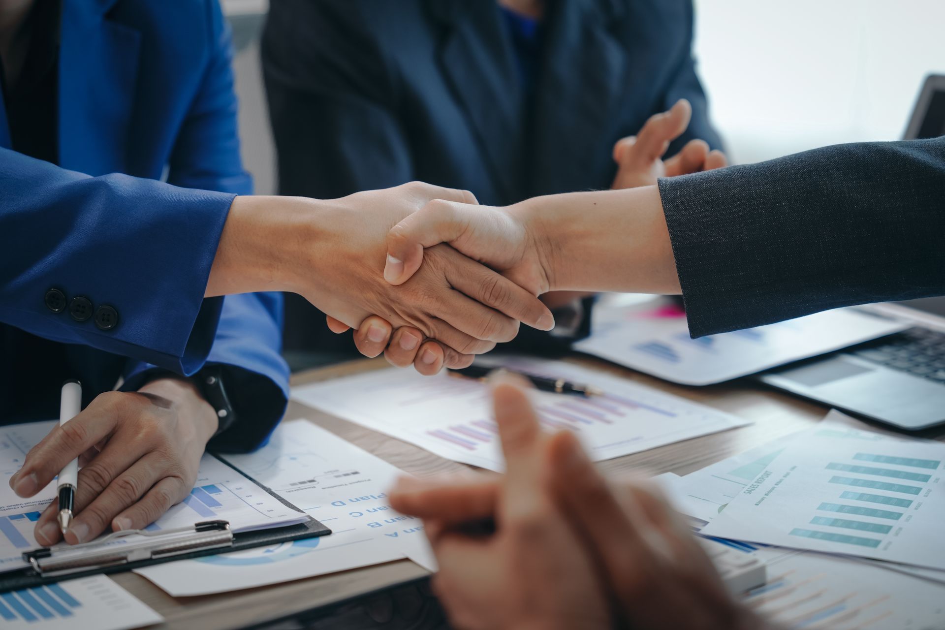 Businesspeople shaking hands over a table with documents and a laptop, symbolizing agreement.