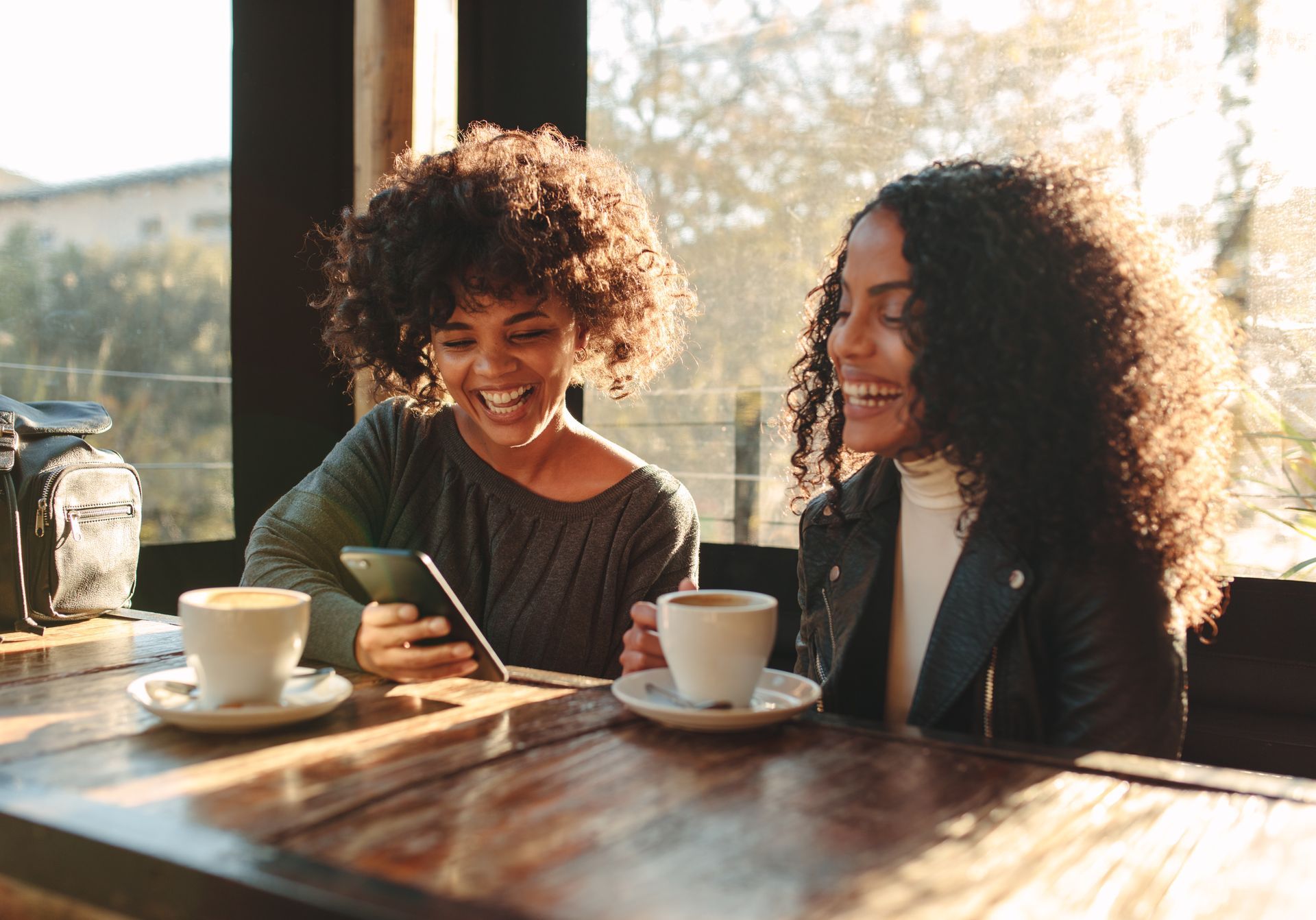 Two people smiling over coffee at a cafe table, one holding a phone. Sunlight streams in.