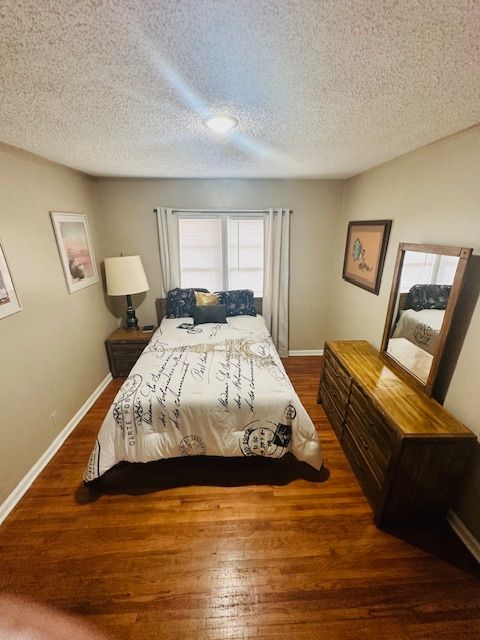 Bedroom with bed, dresser, nightstand, and window with curtains; wood floor and neutral walls.