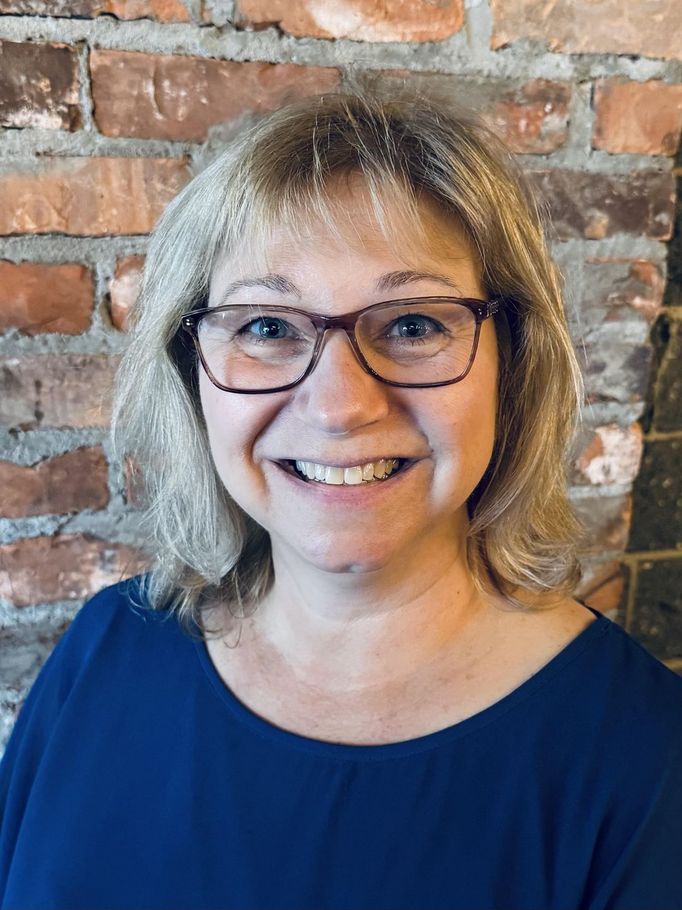 Woman with blonde hair and glasses smiles in front of a brick wall, wearing a blue top.